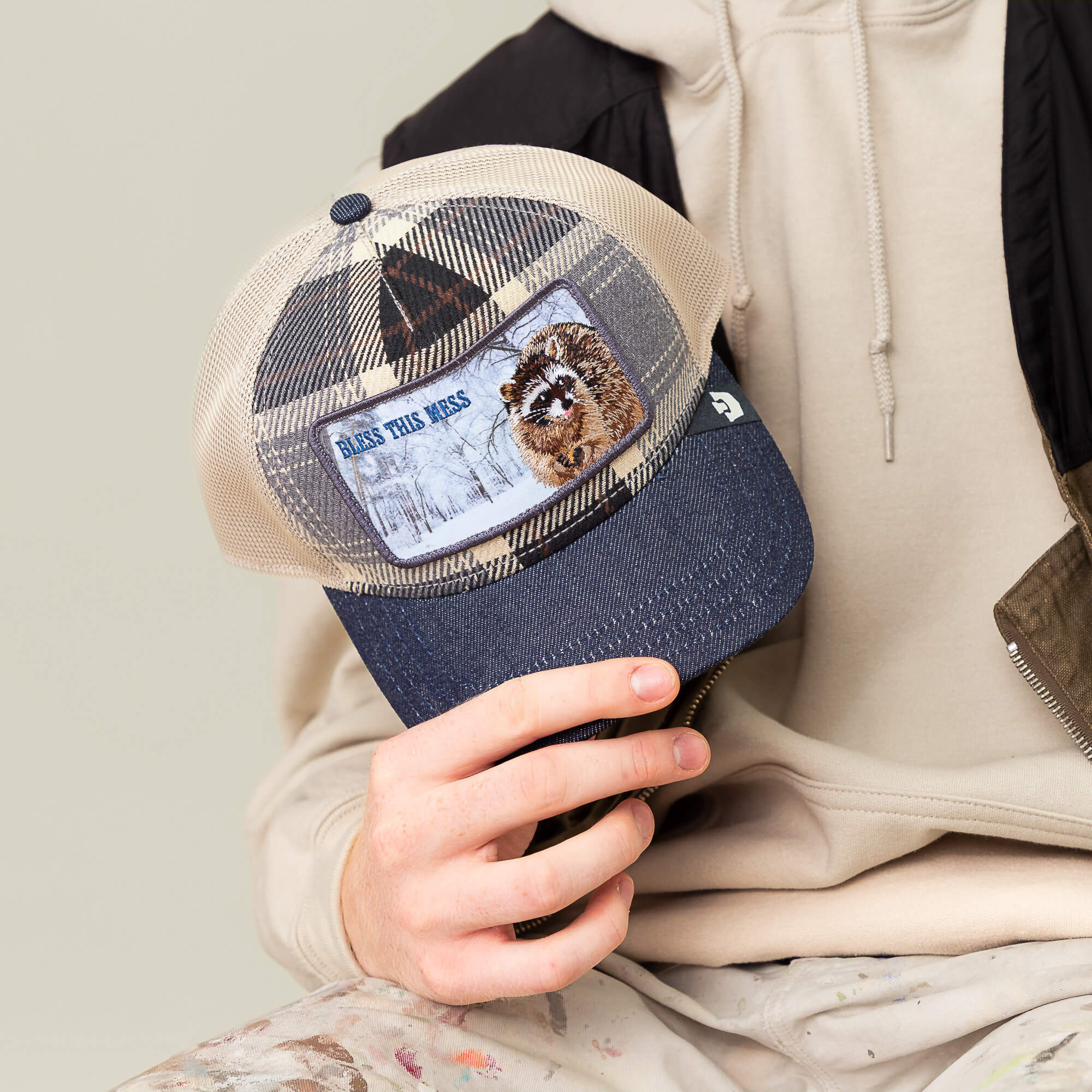 A person in a beige hoodie and paint-splattered pants holds the Goorin Bros. “Free Spirited” plaid crown trucker hat featuring a dog patch and the words “Bless This Mess.”.