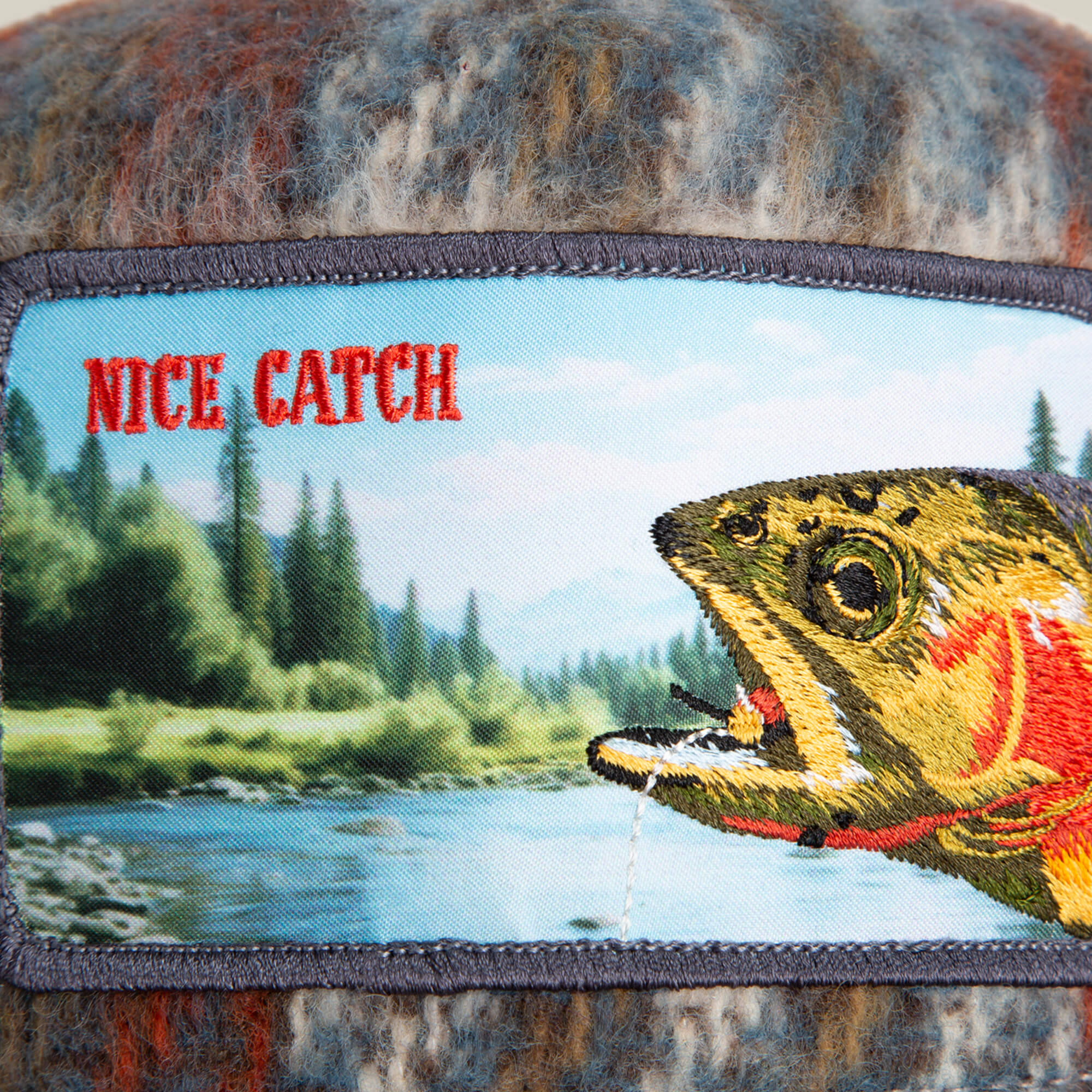 The Down by the River patch by Goorin Bros. features an embroidered fish and river with pine trees and mountains, evoking a cozy flannel fishing trip vibe. "NICE CATCH" stands out in bold red letters.