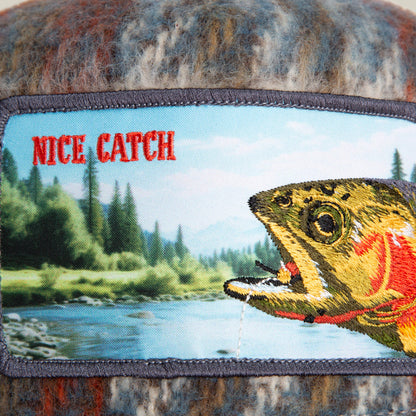 The Down by the River patch by Goorin Bros. features an embroidered fish and river with pine trees and mountains, evoking a cozy flannel fishing trip vibe. "NICE CATCH" stands out in bold red letters.