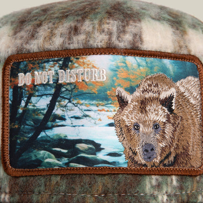 The "Don't Poke the Bear" camo plaid cap by Goorin Bros. features a fabric patch with an embroidered bear, river scene, trees, and "DO NOT DISTURB" text, blending flannel comfort with rugged outdoor style.