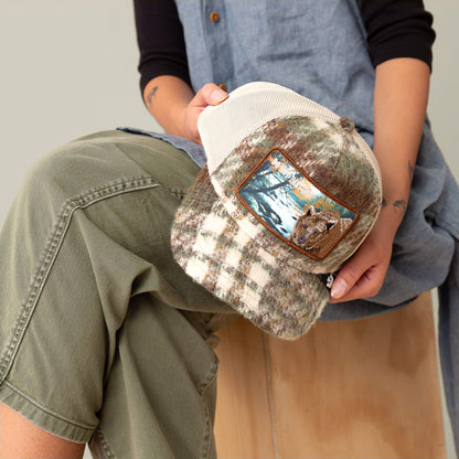 Person holds the Goorin Bros. "Don't Poke the Bear" cap, featuring camo plaid, an outdoor scene, and a bear patch—pairing flannel comfort with a playful warning. Worn with green pants and a blue shirt.
