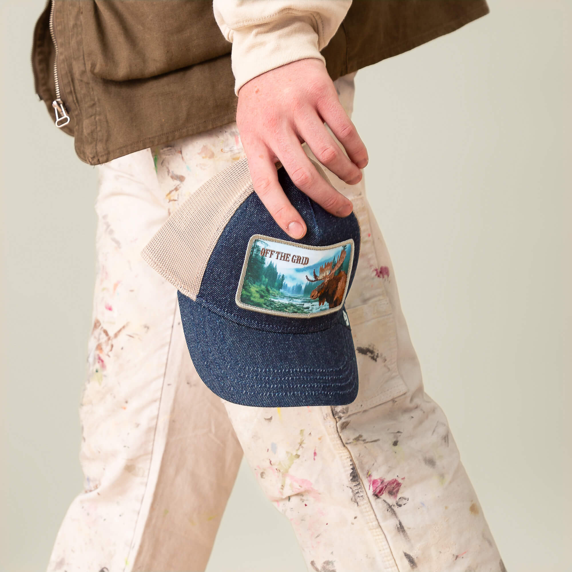 A person in paint-splattered pants holds the Goorin Bros. Staycation cap, featuring a denim crown, blue mesh, and an Off The Grid patch with a mountain nature scene.