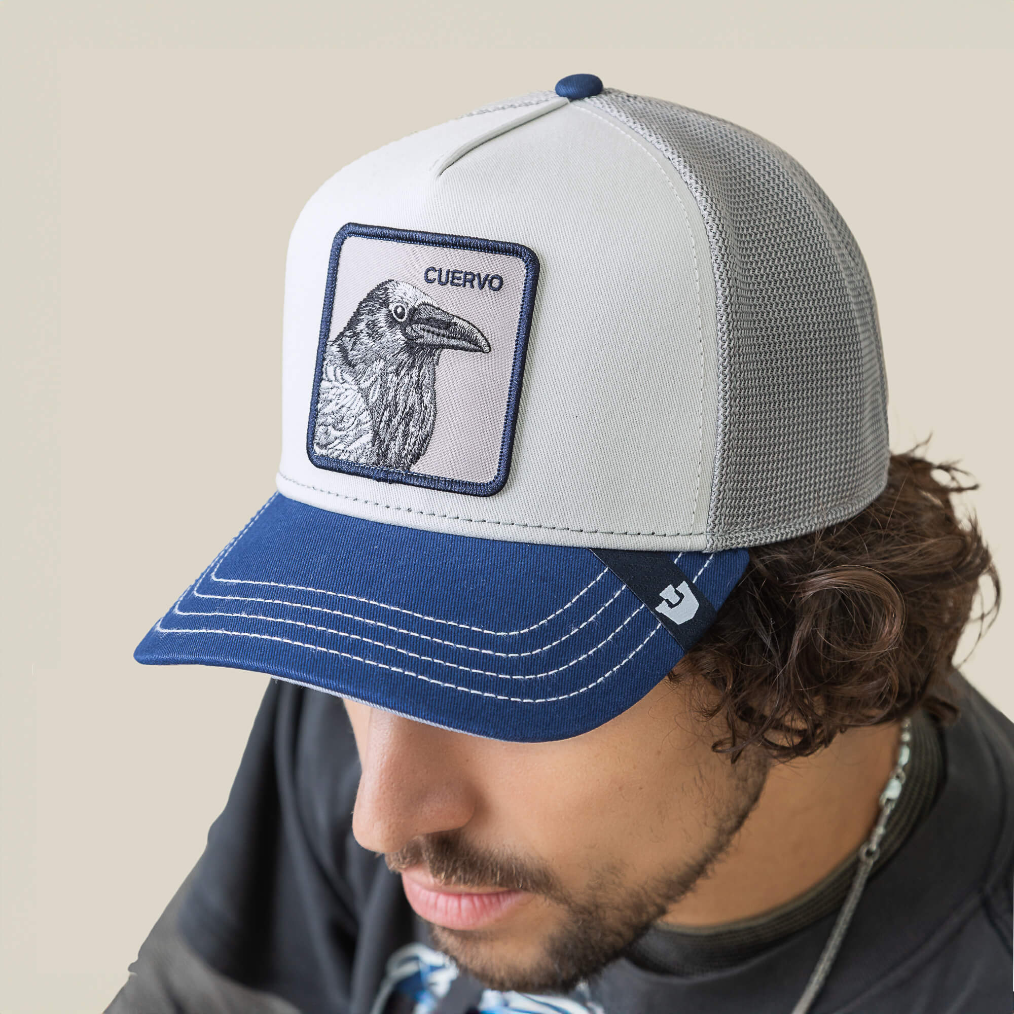 Man wearing a Goorin Bros. Tricolor Raven mesh trucker hat with white and blue panels, a navy bill, and a raven patch labeled CUERVO.