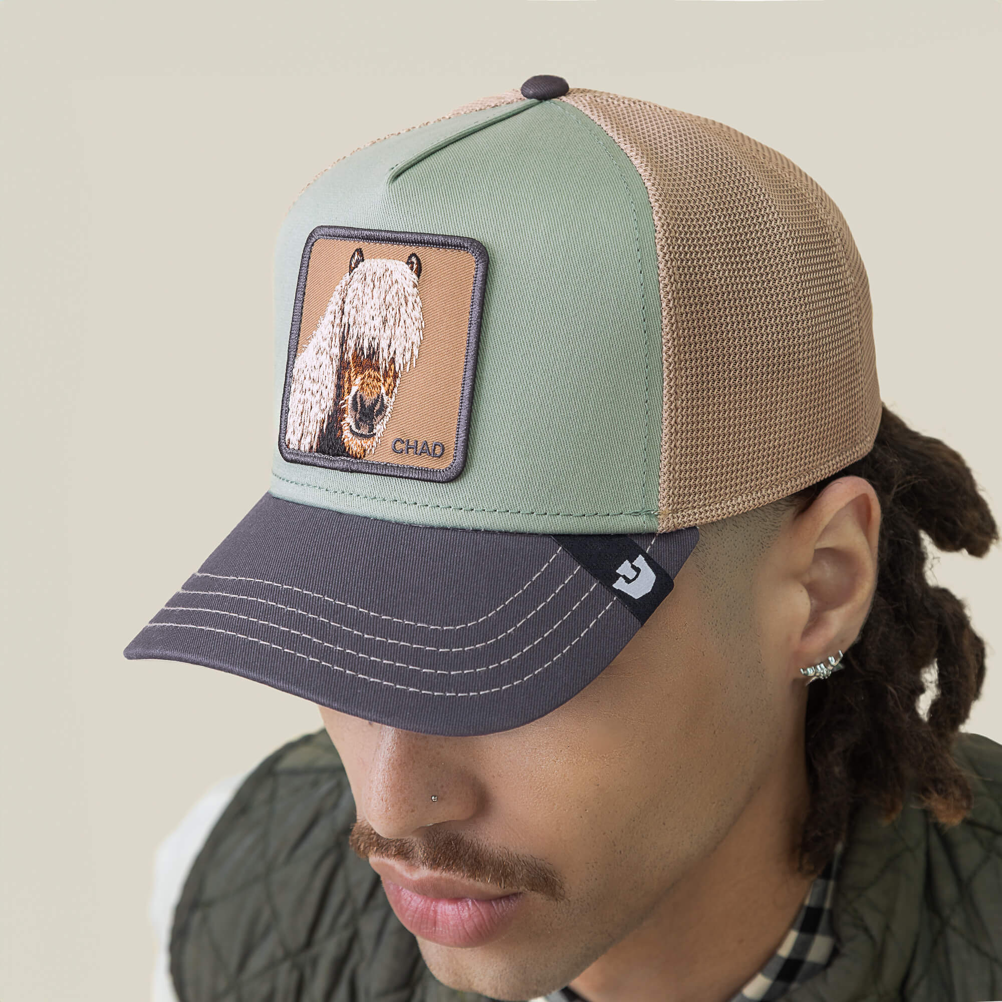 A person wearing the Goorin Bros. Tricolor Horse hat, featuring mint green fringe, a mustache, earrings, and a patch with an illustrated animal and the name CHAD.
