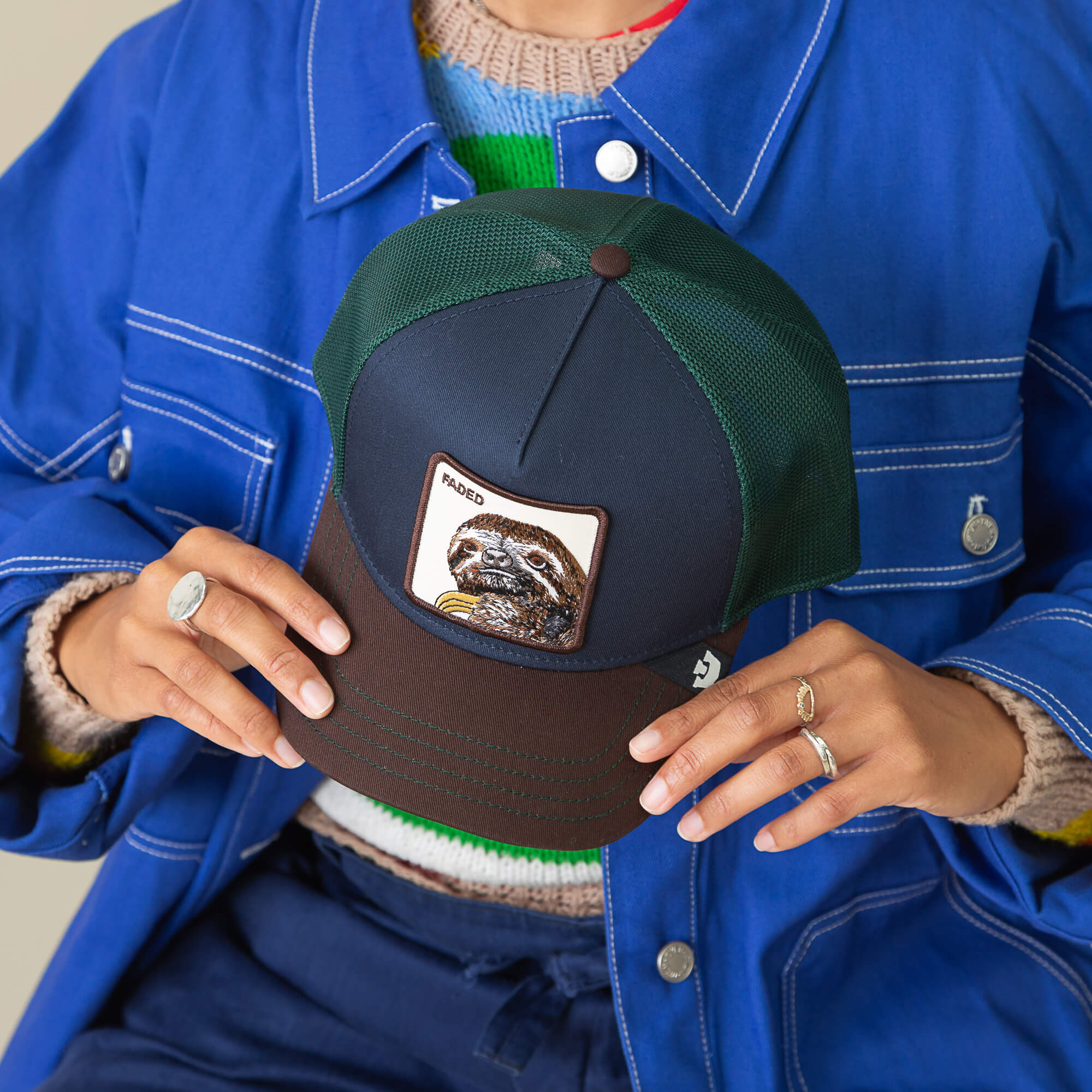 Person holding the Goorin Bros. Tricolor Sloth hat—a stylish navy mesh cap with a sloth patch—while wearing a blue jacket and multiple rings; a standout fashion choice.