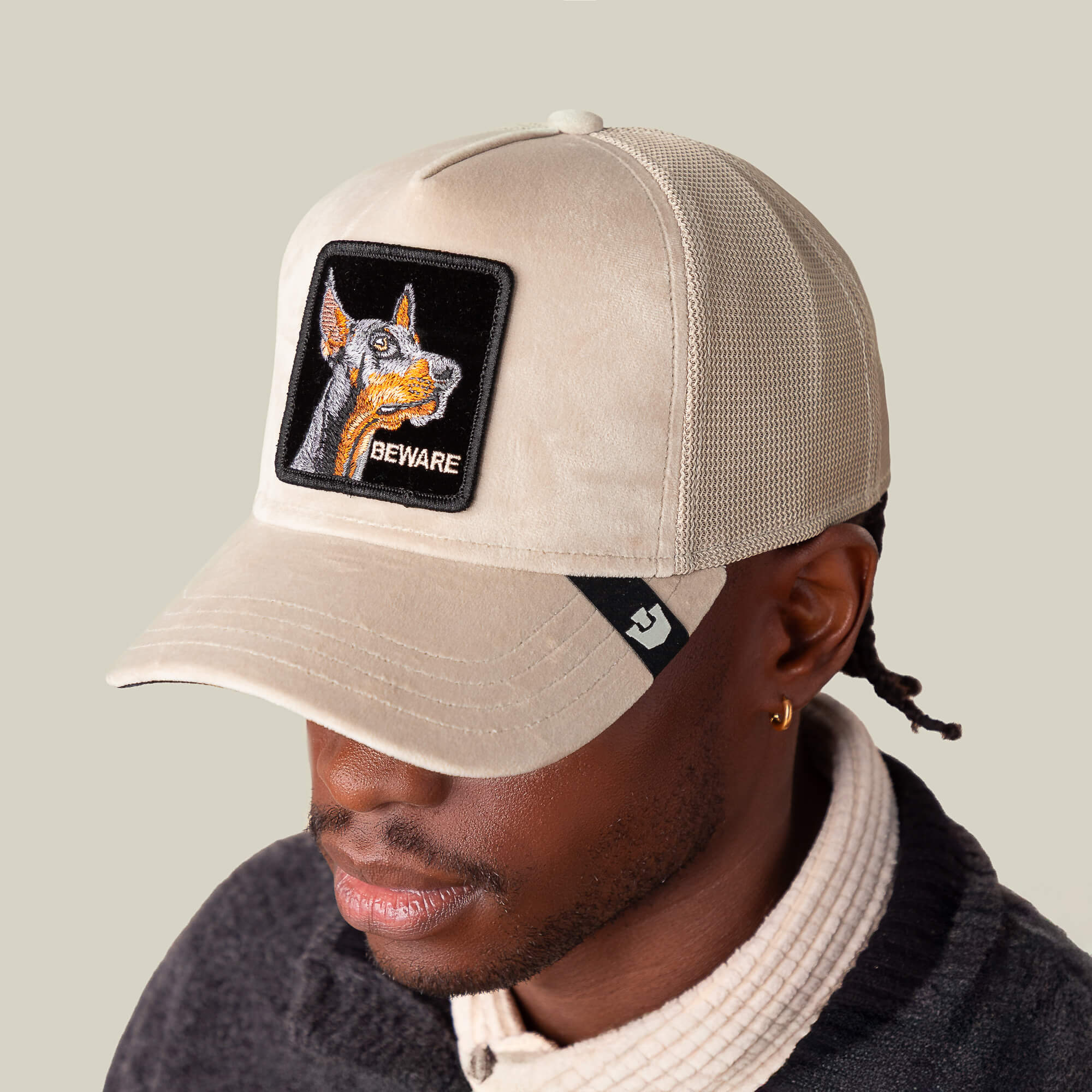 A person wears the Goorin Bros. Velour Doberman cap—a luxury beige hat featuring a patch with a Doberman’s head and "BEWARE" on the front—while facing slightly downward.