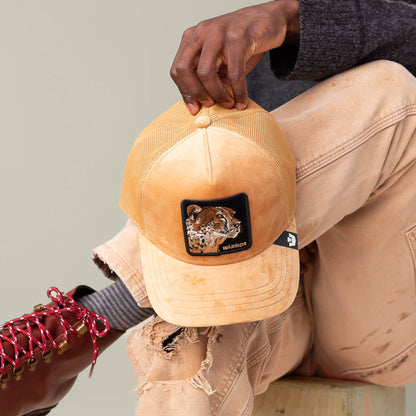 A person in tan pants and red boots holds the Goorin Bros. Velour Cheetah tan mesh baseball cap featuring a jaguar face patch and the word "WARRIOR" embroidered on it.