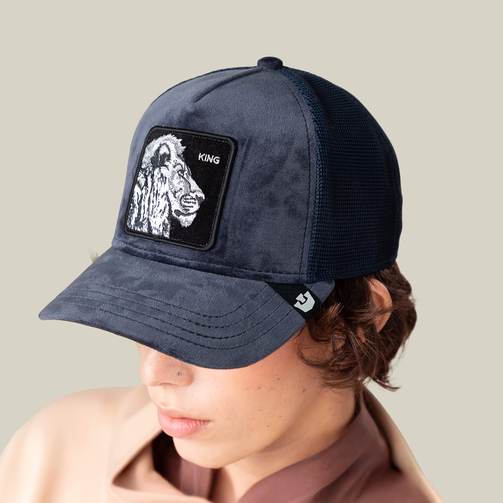 A person in a dark blue Goorin Bros. Velour Lion hat with a KING patch looks downward, wearing a beige top against a plain background.