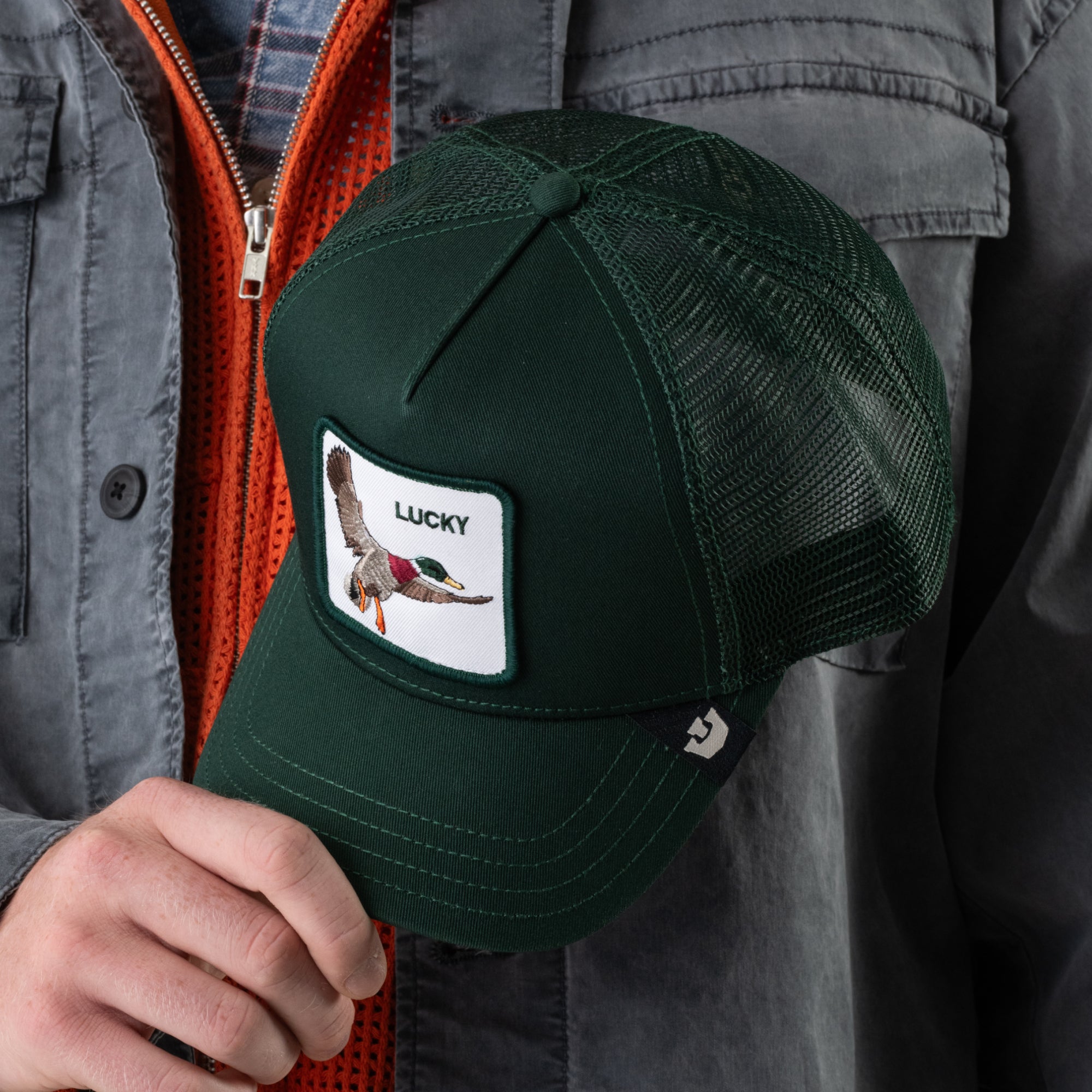A person wearing a gray jacket with an orange zipper holds Goorin Bros.’ “The Lucky Duck”—a dark green mesh trucker cap featuring a charm patch with a flying duck and the word “Lucky.”.