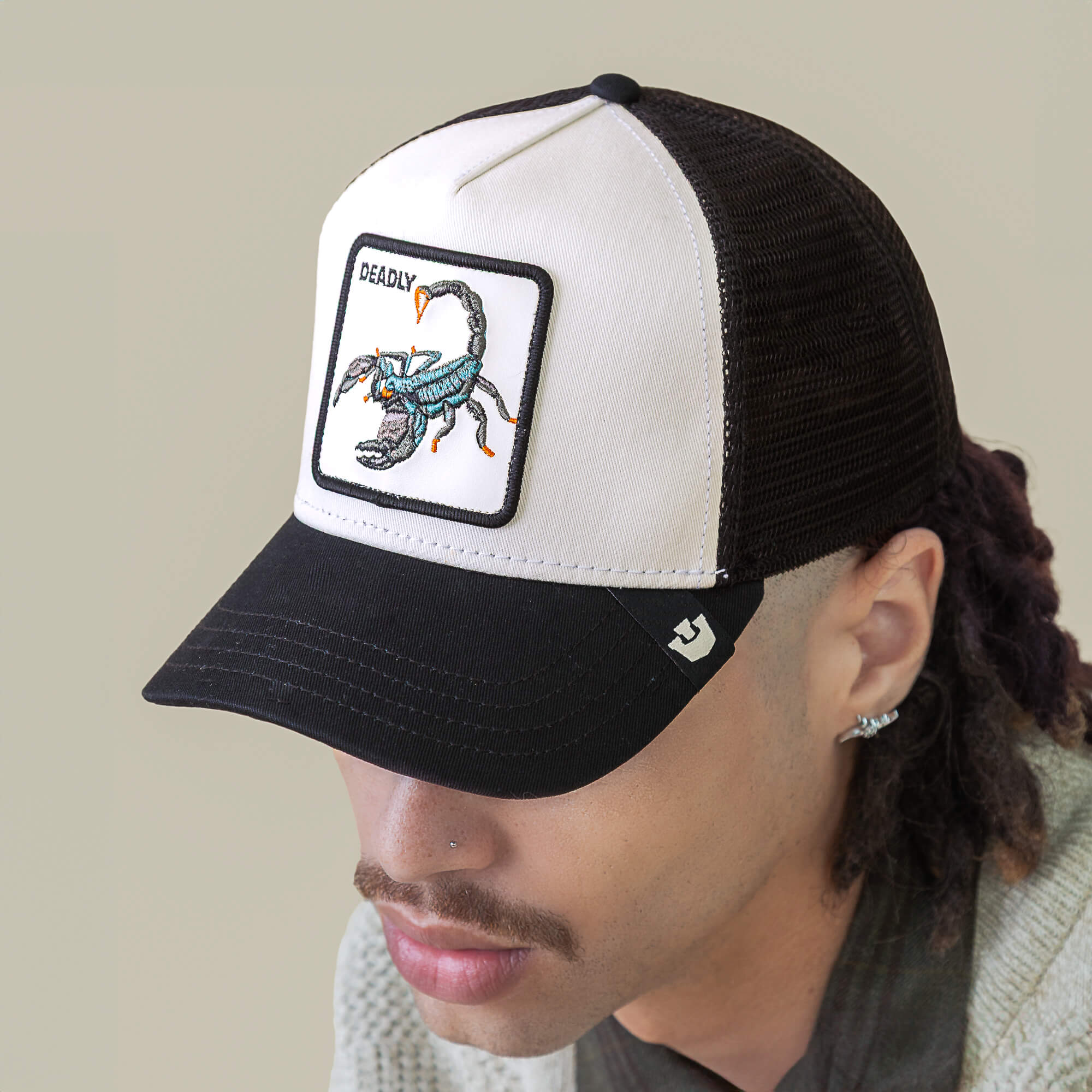 A person wears the "Deadly" black and white mesh hat by Goorin Bros., featuring a front scorpion patch. The person's face is partially visible.