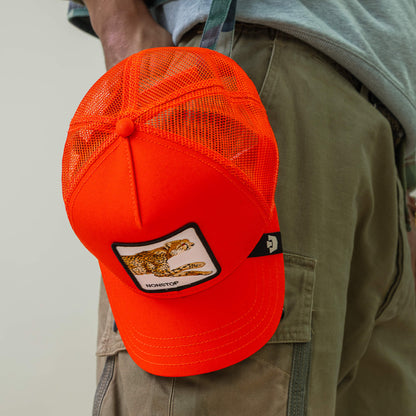 A bright orange Goorin Bros. "The Nonstop Cheetah" mesh trucker cap with cheetah patch hangs from the side pocket of green cargo pants, injecting a fast-paced vibe into the outfit.
