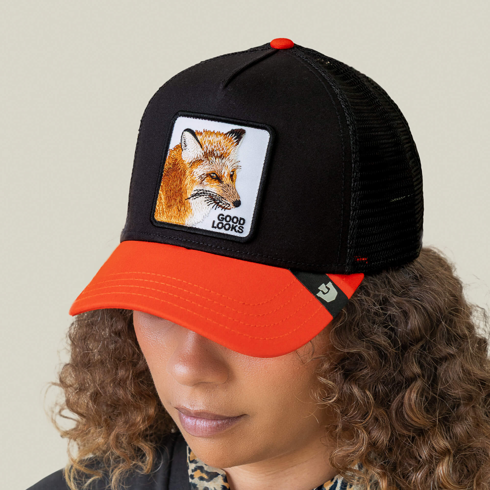 Person wearing the Goorin Bros. Good Looks Fox trucker cap featuring a black and orange design with a fox patch and "GOOD LOOKS" on the front, exuding a stylish runway-ready vibe while looking downward with curly hair.