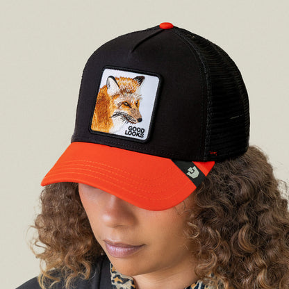 Person wearing the Goorin Bros. Good Looks Fox trucker cap featuring a black and orange design with a fox patch and "GOOD LOOKS" on the front, exuding a stylish runway-ready vibe while looking downward with curly hair.