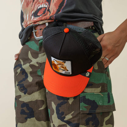 Wearing camouflage cargo shorts and a graphic tee, someone hooks the Goorin Bros. Good Looks Fox trucker hat—featuring an orange and black fox patch—to their belt, merging farmyard chic with effortless style.