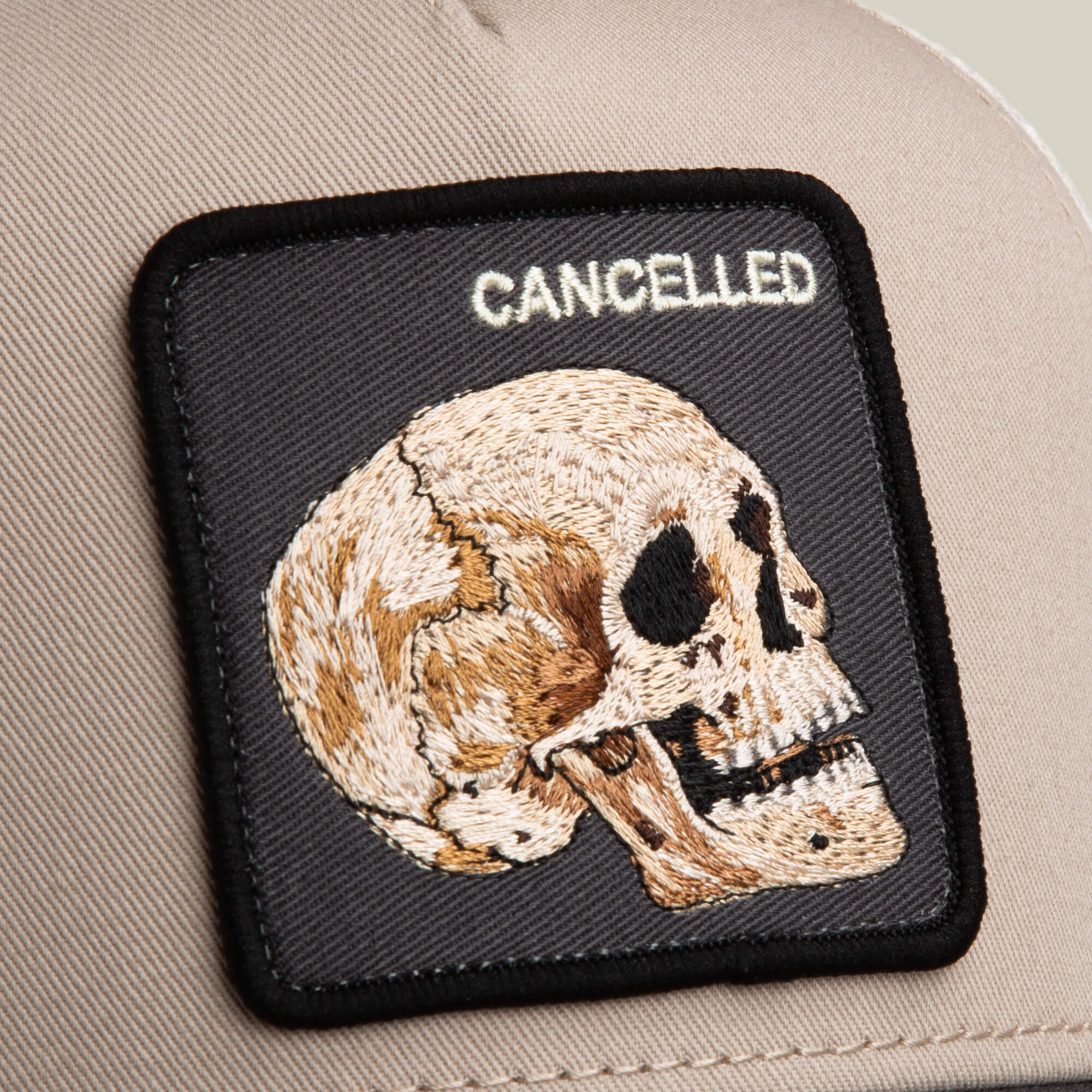 A close-up of The Cancelled Skull trucker hat by Goorin Bros., featuring a beige design with a stitched skull patch and "CANCELLED" in white, perfect for anyone who likes to speak their mind.