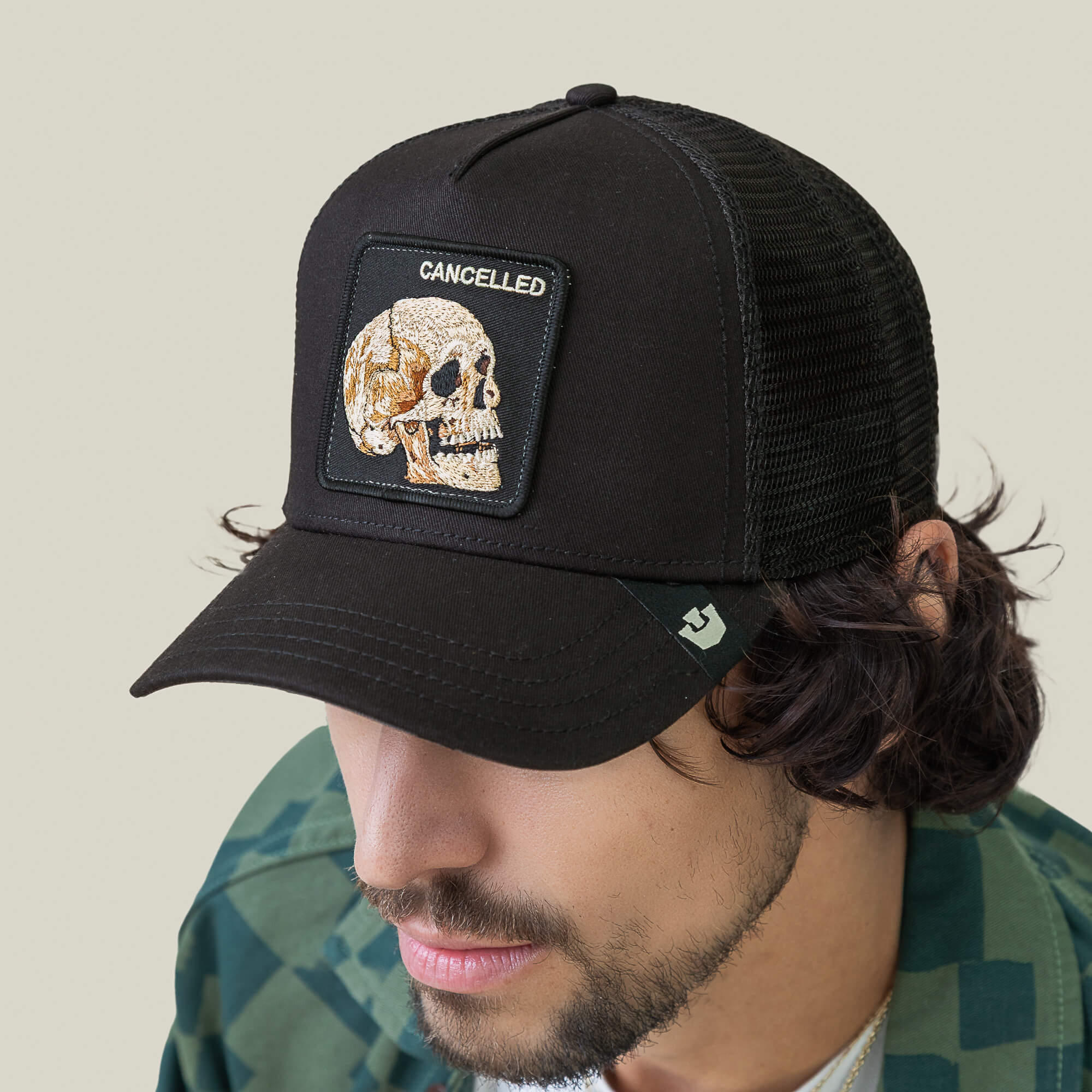 A man in a green plaid shirt looks downward, wearing Goorin Bros.' The Cancelled Skull black mesh trucker hat with a stitched skull patch and CANCELLED text.
