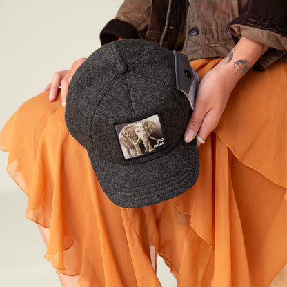 A person in an orange skirt holds Goorin Bros.’ Kind of trucker beanie in dark gray, featuring an elephant patch and "BIG DEAL" text, crafted from the ultimate feel-estate for comfort.
