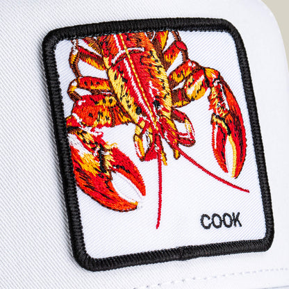 Close-up of a white patch with an embroidered red lobster and "COOK" stitched at the corner, featured on the Lemme Cook trucker hat by Goorin Bros., showcasing a stylish mesh back design.