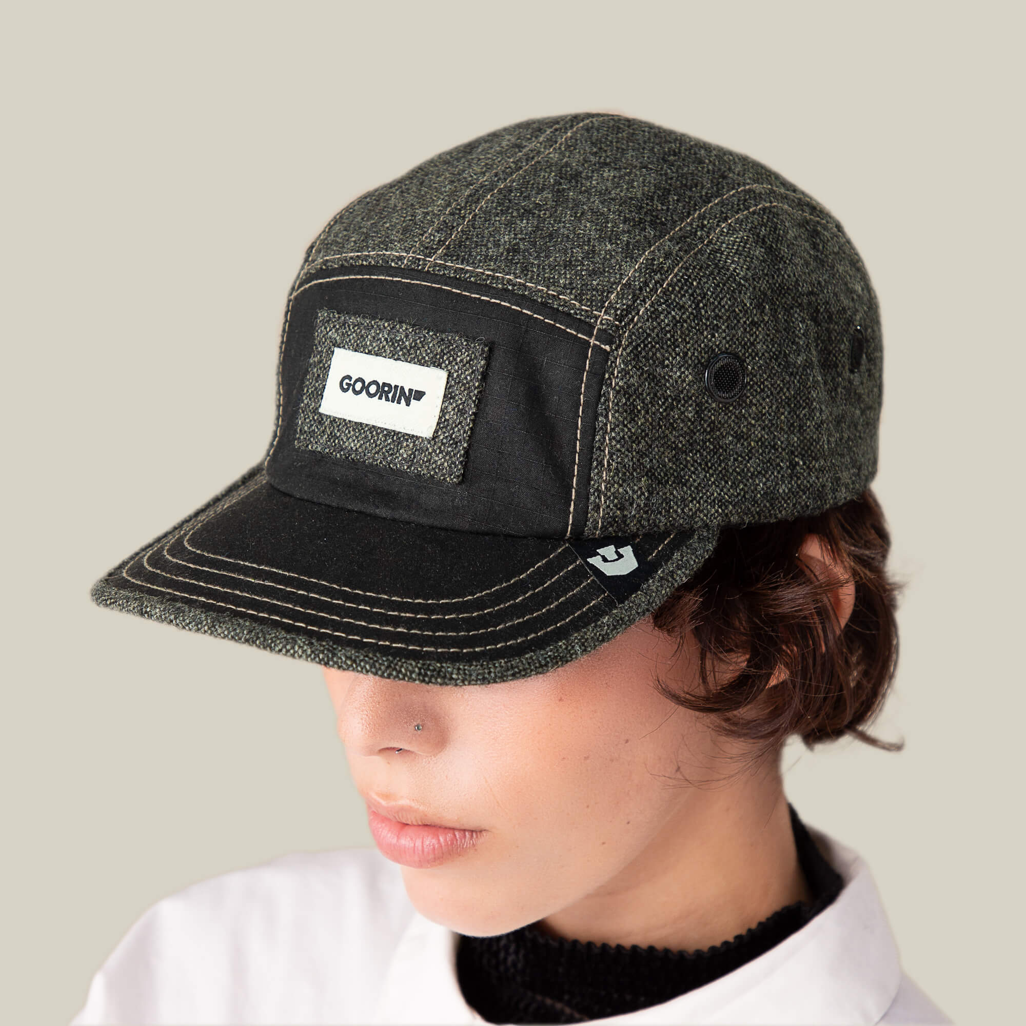 A person wearing the Goorin Bros. Billed Demand cap in dark grey and black with a flat brim and signature patch, looking slightly downward against a plain light background.