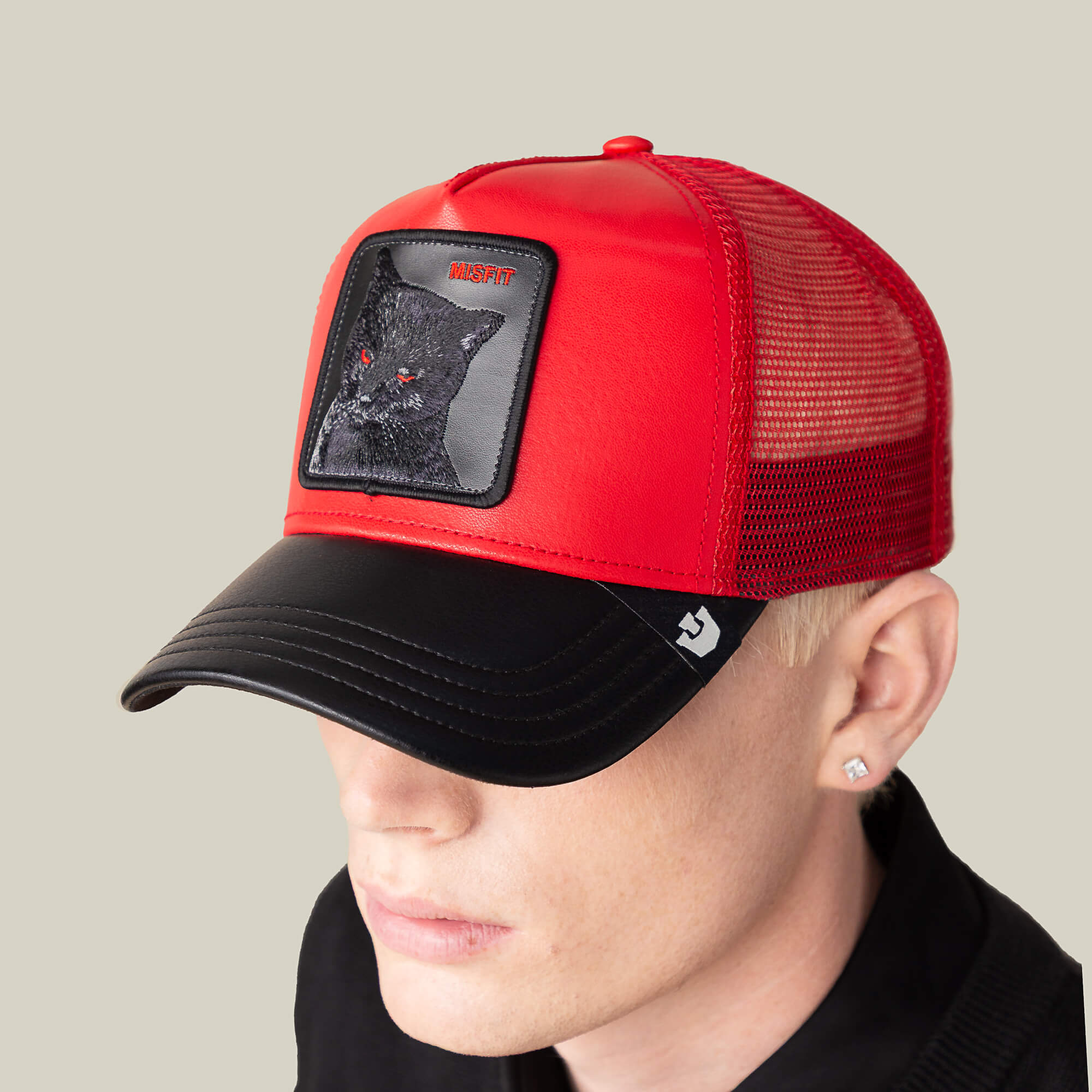 A person in a Goorin Bros. Courtside Misfit red faux leather hat with a black cat patch, black shirt, and stud earring stands against a plain background, embodying the signature Courtside Misfit look.