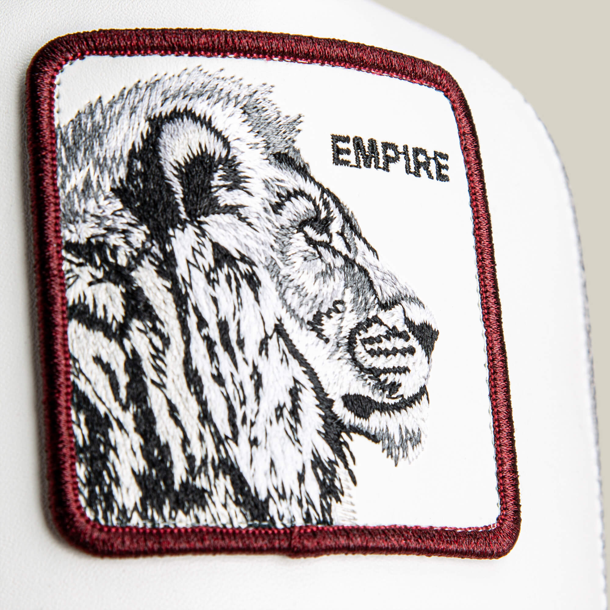 Courtside Empire by Goorin Bros.: a rectangular patch featuring a black and white embroidered lion head with a leather-look crown, "EMPIRE" in all caps, maroon border stitching, and white background—ideal for adding courtside or sneaker style.