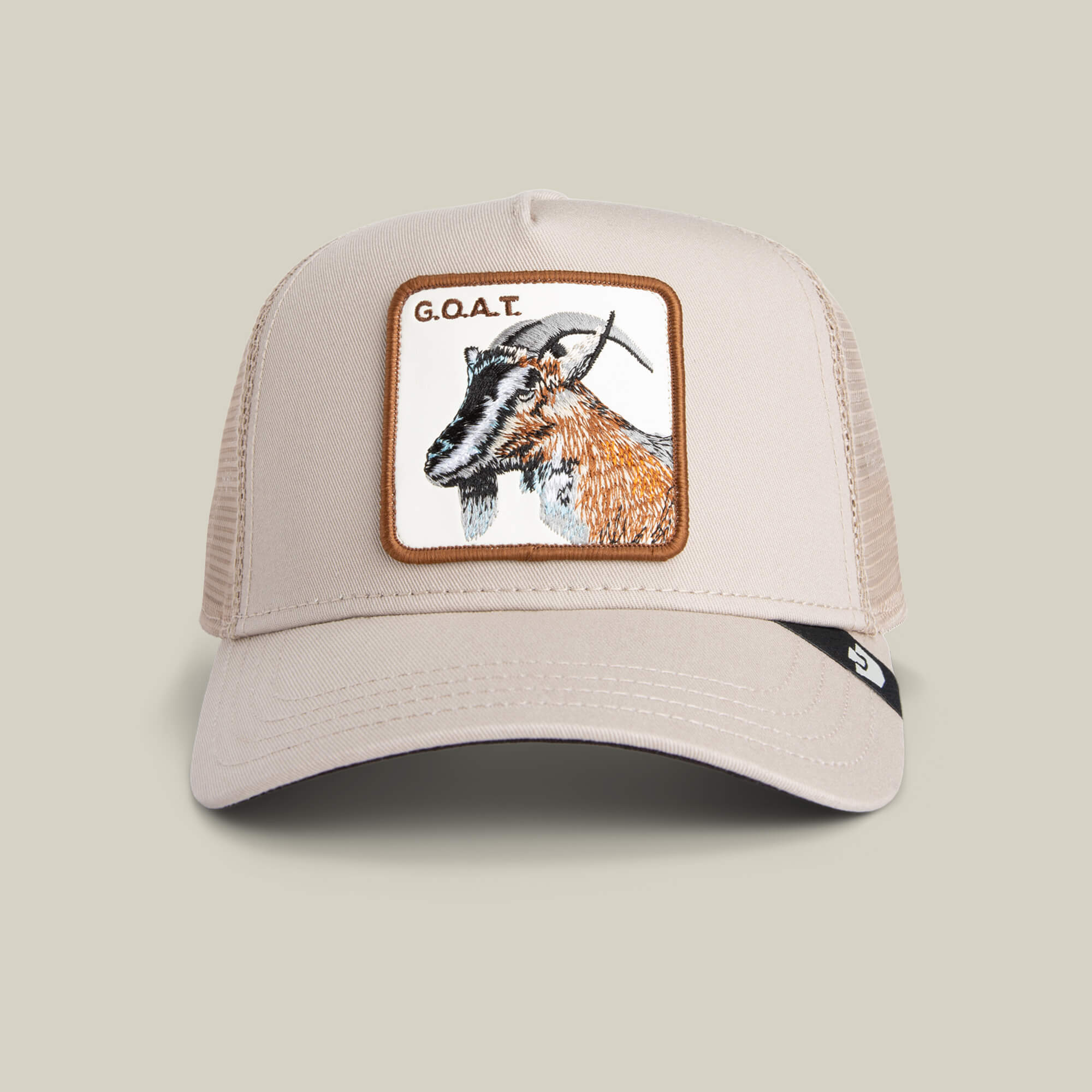 The G.O.A.T by Goorin Bros. is a beige mesh trucker hat in neutral tones with a bold front patch featuring an embroidered goat and the text "G.O.A.T.
