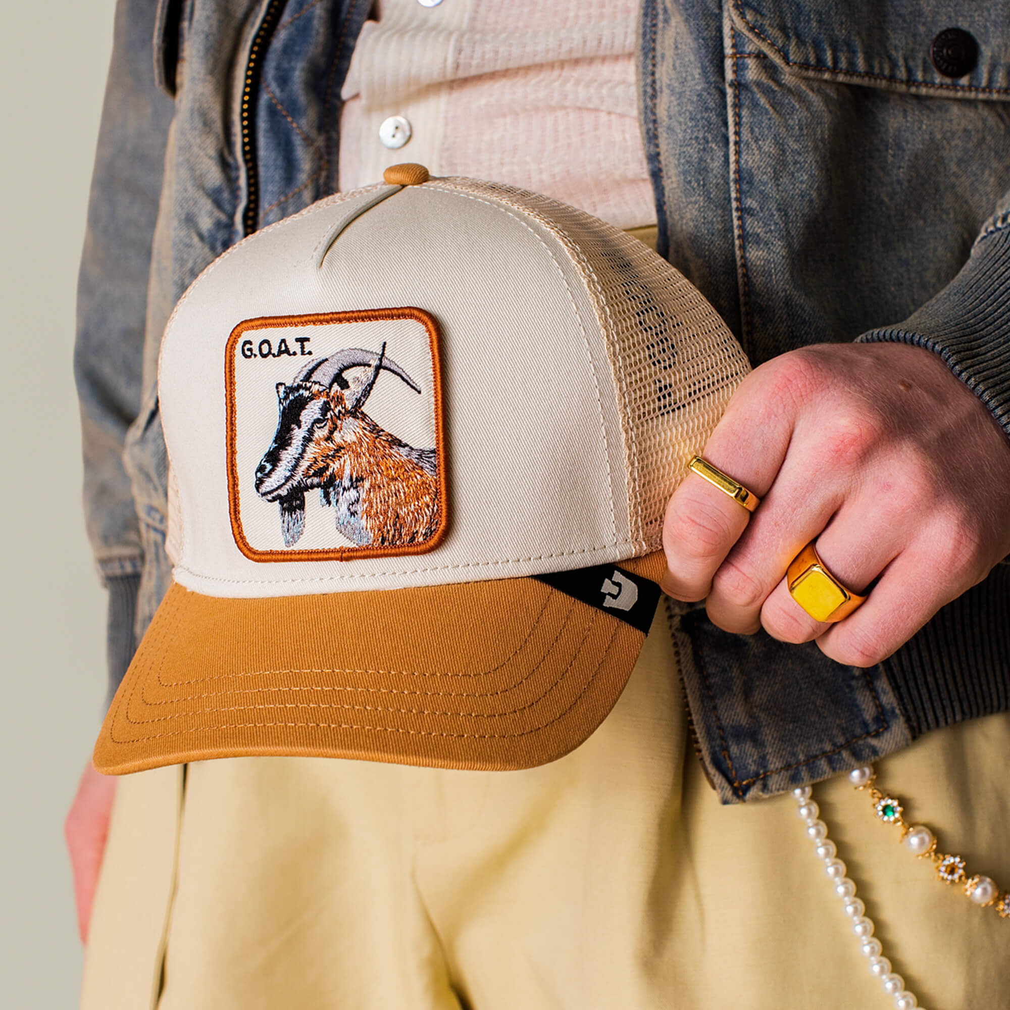 Wearing beige pants and a denim jacket, a person holds Goorin Bros.' The G.O.A.T hat in neutral tones, featuring a bold patch with an embroidered goat and "G.O.A.T" label, accessorized with gold rings and a beaded chain.