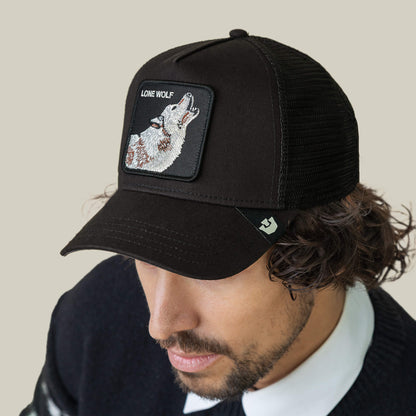 A man wears The Lone Wolf cap by Goorin Bros., featuring a black mesh trucker style and "Lone Wolf" patch with a howling wolf, exuding outlaw energy against a plain beige background.