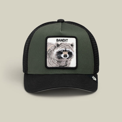 The Goorin Bros. Bandit Trucker is an olive green and black colorblock trucker hat with a mesh back and snapback closure, featuring a rectangular patch with a detailed raccoon illustration and the word Bandit on the front panel.
