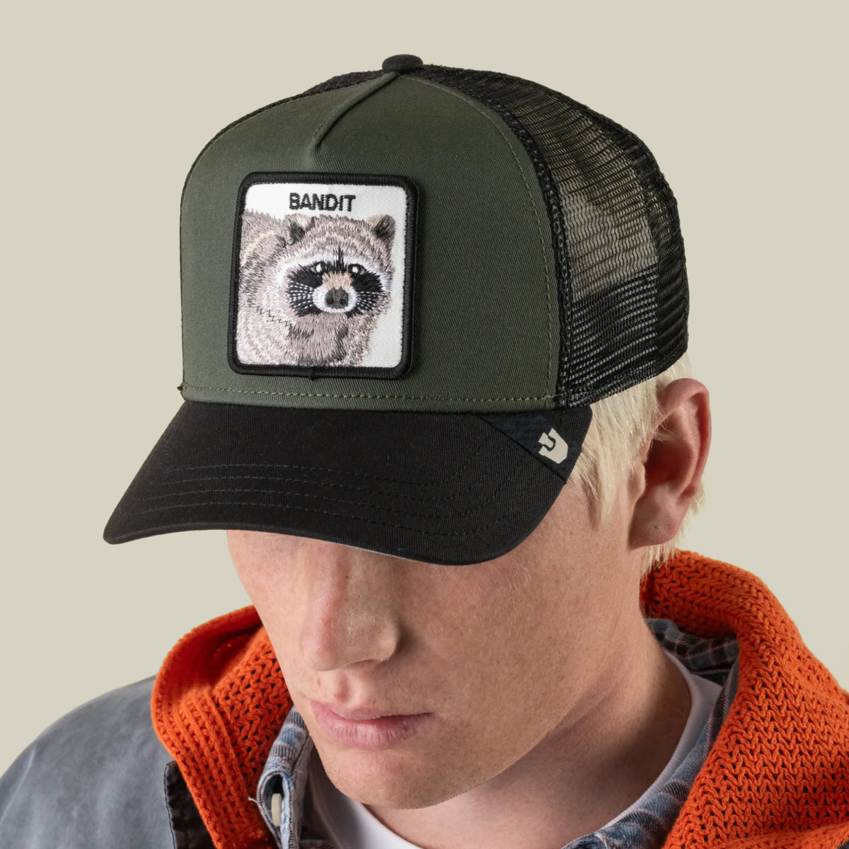 A person wearing a Goorin Bros. Bandit Trucker cap in green and black, featuring a raccoon insignia patch labeled “BANDIT” on the front, along with an orange hoodie and a plaid shirt.