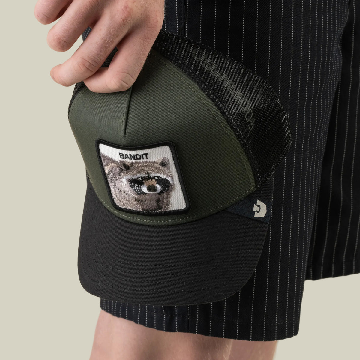 A person is holding a green and black Goorin Bros. Bandit Trucker hat with a mesh back, featuring a BANDIT raccoon patch, while wearing black pinstriped shorts.