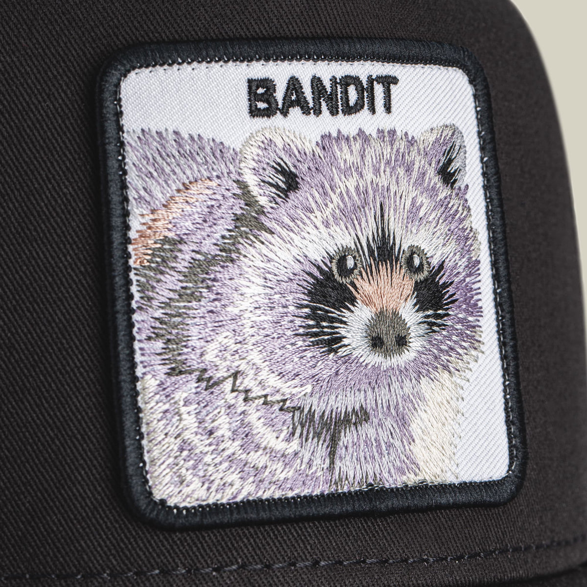 The Goorin Bros. Bandit Trucker is a black trucker hat featuring a detailed embroidered raccoon patch on the front, shown in close-up detail against the dark fabric.