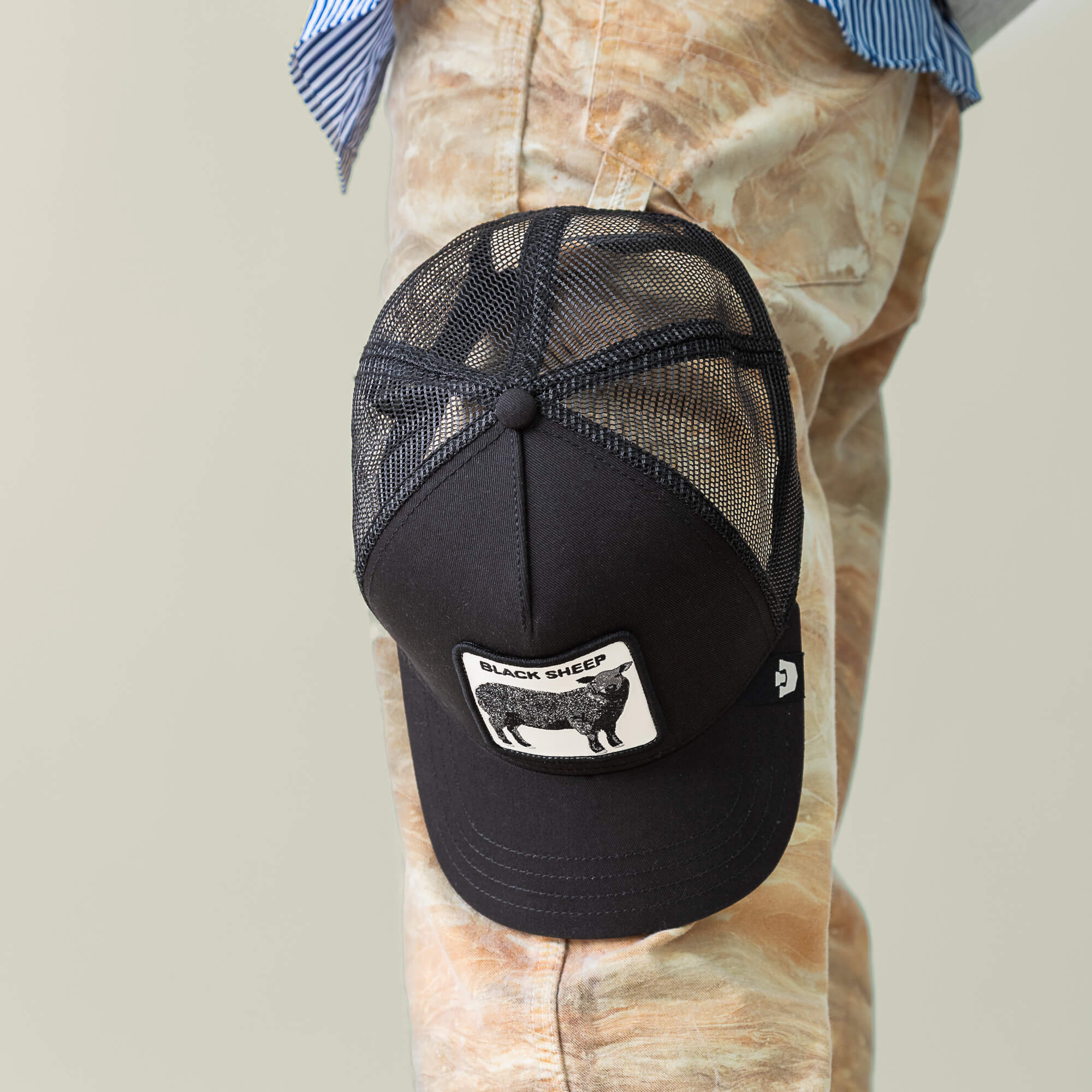 The Black Sheep by Goorin Bros.—a black mesh trucker hat with a signature patch—rests on someone's knee. They pair it with marbled tan pants and a blue-and-white striped shirt for a standout look.