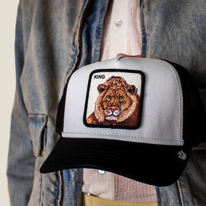 A person in a denim jacket holds the King Trucker cap by Goorin Bros., a black and white baseball hat featuring a lion patch with the word "KING"—a bold nod to power dressing.
