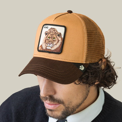 A man wears the Goorin Bros. King Trucker, a brown and tan mesh hat featuring a KING lion patch on the front, and looks down against a plain beige background.