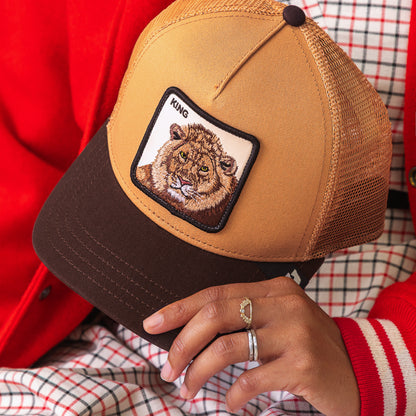 Wearing rings and dressed in power style with a red jacket and checkered clothes, they confidently hold the Goorin Bros. King Trucker cap—a brown and tan mesh hat featuring a bold lion patch labeled KING.