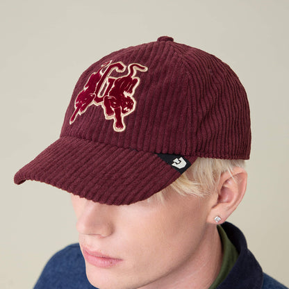 A person wearing the Goorin Bros. Bleacher Report Papa Cap, featuring a burgundy corduroy crown, two embroidered cats on the front, a small black tag on the brim, and a stud earring.