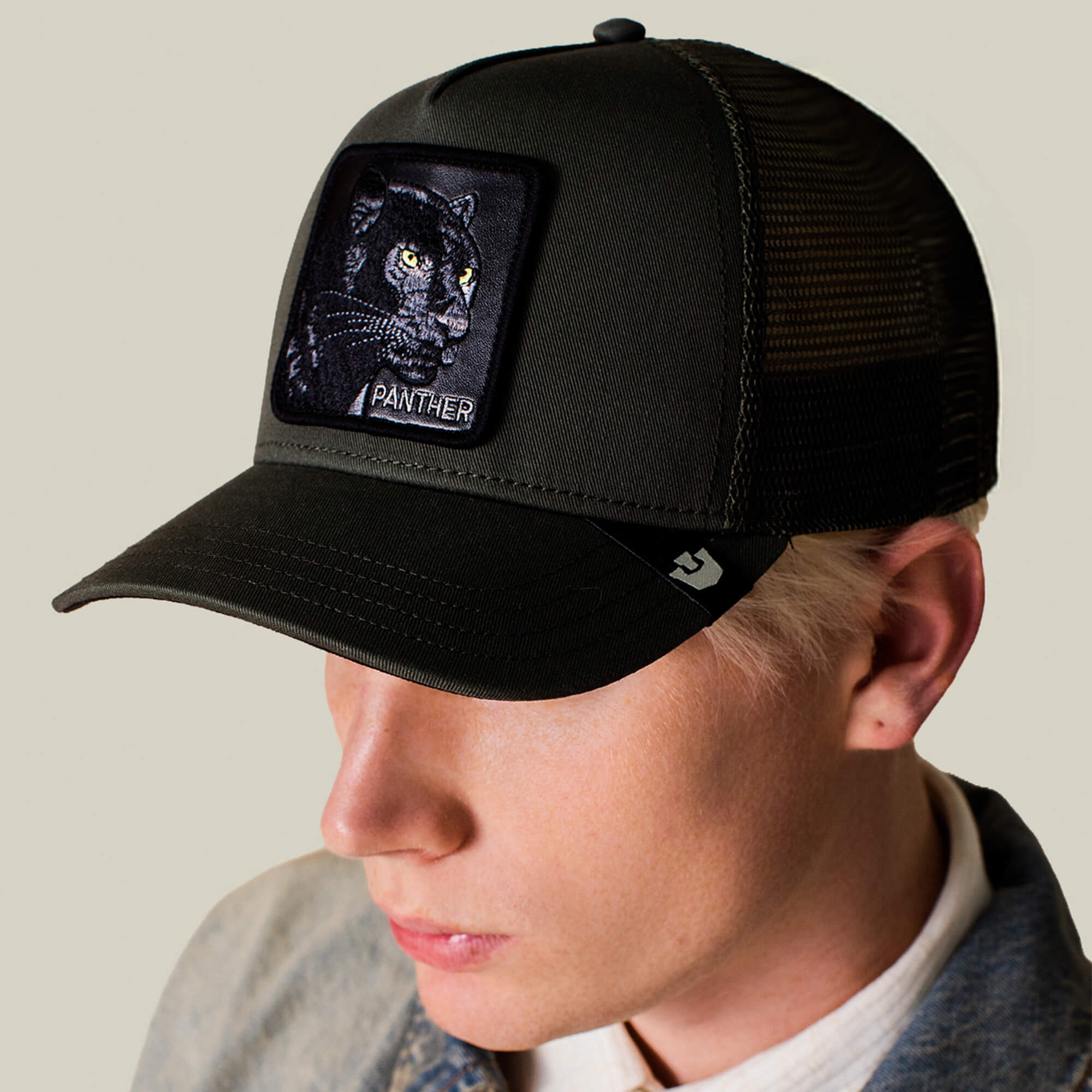 A person wears The Panther by Goorin Bros.—a black mesh trucker hat featuring an embroidered panther patch—styled with a light jacket and a white collared shirt.