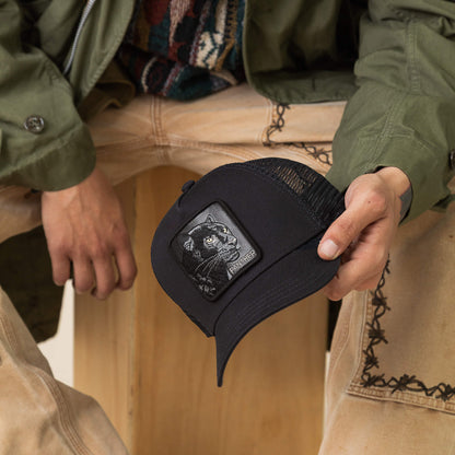 A person sits on a wooden crate, holding Goorin Bros.' The Panther hat featuring a black cat patch, and wearing tan embroidered pants with a green jacket.