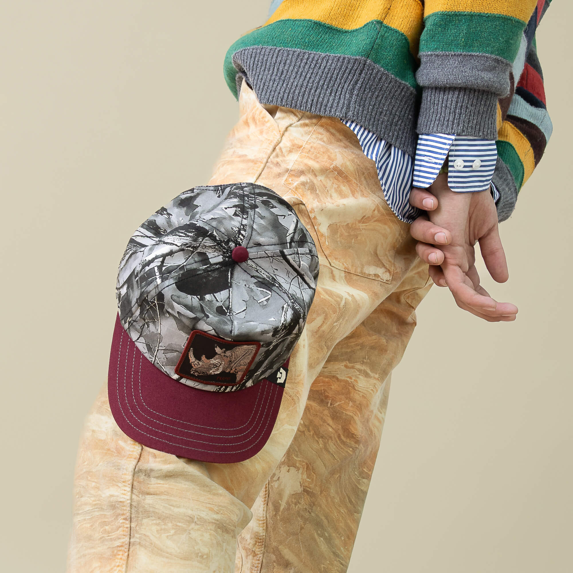 Wearing marbled beige pants, a striped green and yellow sweater, and the Goorin Bros. Tuff Camo patterned low crown cap clipped to their side, they rest their hand near their hip.