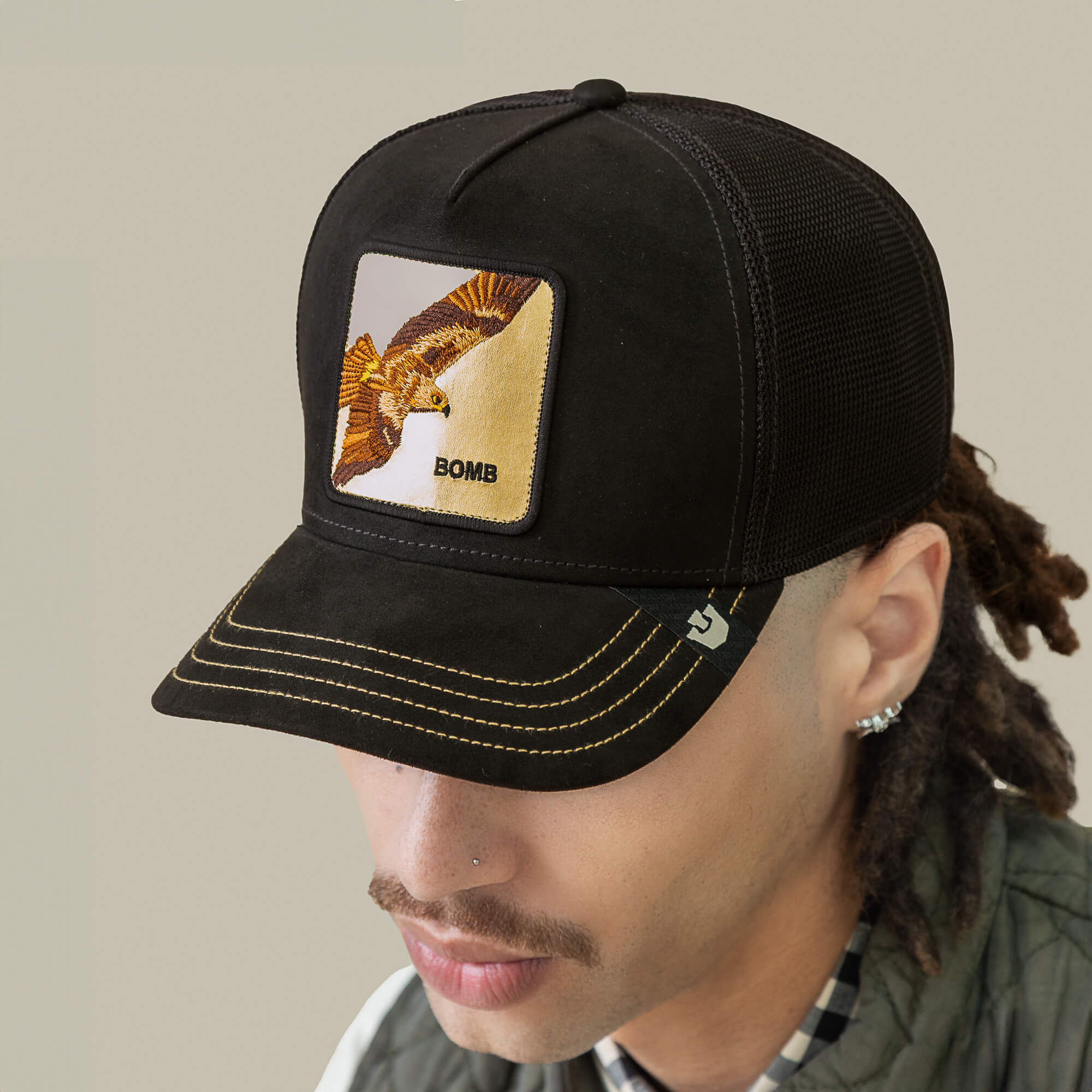 A person wears a black mesh Goorin Bros. trucker hat with yellow stitching and a patch showing a bird and the words "Golden Bomb," inspiring you to rise high.