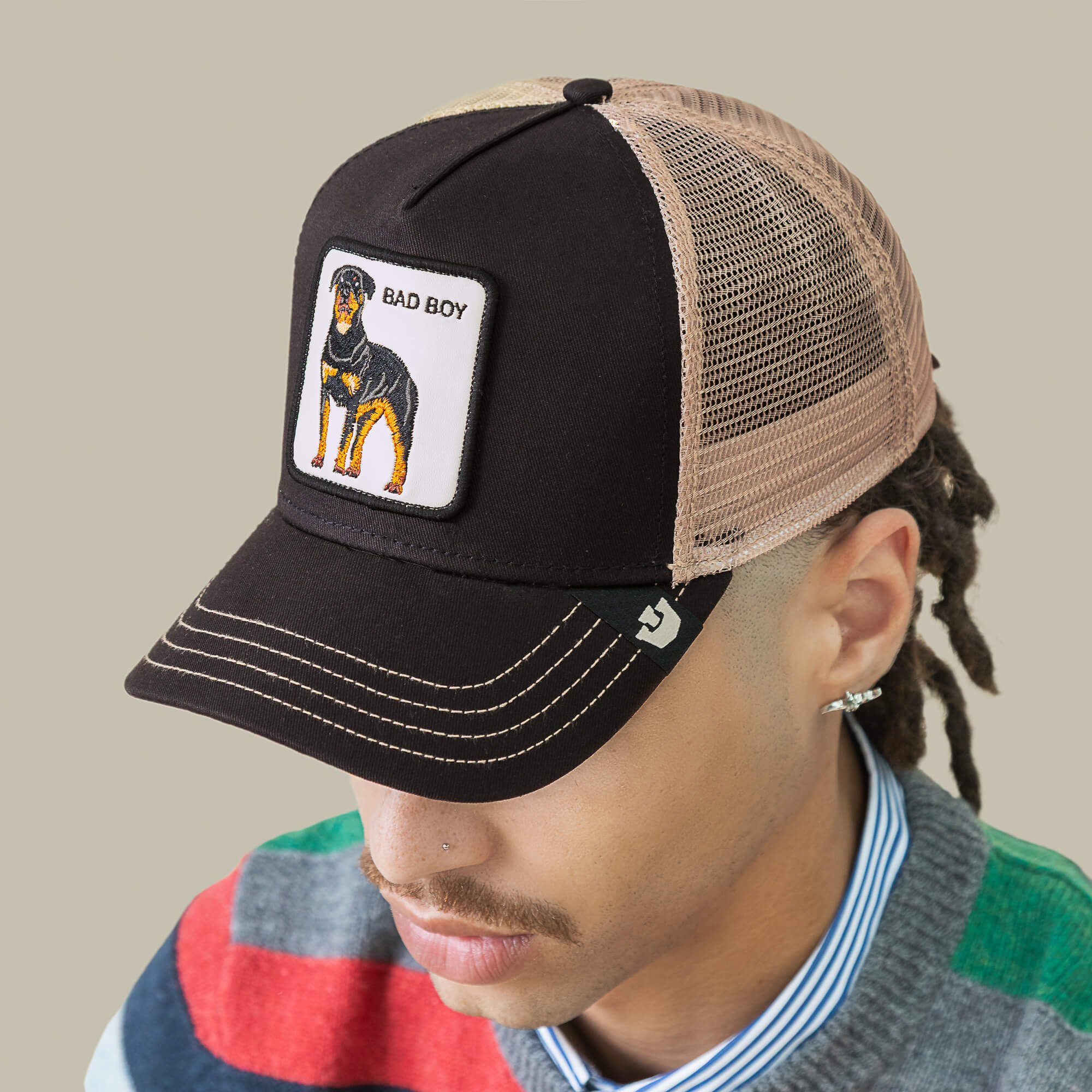Person wearing The Bad Boy mesh trucker hat by Goorin Bros.—a black and tan cap with a dog patch—styled with a striped shirt and colorful sweater, making this bold accessory the standout piece of the outfit.