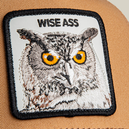 A close-up of the Wise Ass Trucker cap by Goorin Bros., featuring an embroidered owl patch with yellow eyes and "WISE ASS" above it, sewn onto a tan crown.