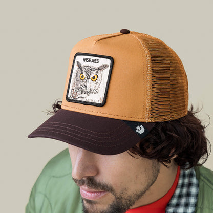 A man wearing a Goorin Bros. Wise Ass Trucker cap, featuring a brown and tan mesh design with an owl patch, along with a green jacket and red shirt.