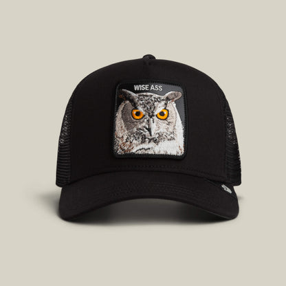 The Wise Ass Trucker by Goorin Bros. is a black mesh cap featuring an embroidered owl patch with orange eyes and the words "WISE ASS" above it.