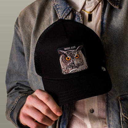 A person in a denim jacket holds the Wise Ass Trucker by Goorin Bros.—a black cap with an embroidered owl and "WISE ASS" text.