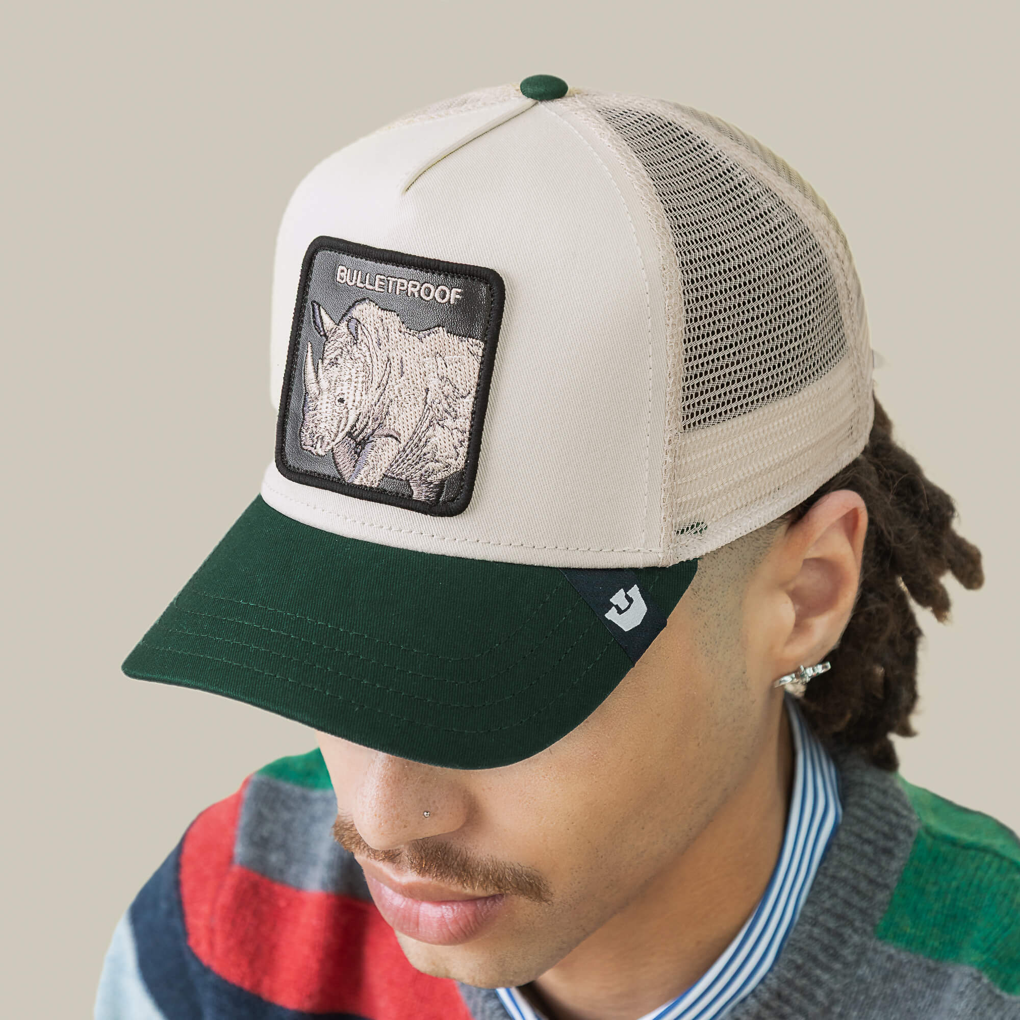 A person wearing the Goorin Bros. Bulletproof Trucker hat, featuring a beige and green design with a rhino patch, styled with a striped shirt and a vibrant sweater that suggests a marshmallow-soft side beneath their Rhino armor.