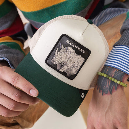 A person with a tattooed arm wears a beaded bracelet and striped sweater, holding the Goorin Bros. Bulletproof Trucker hat—beige and green with a rhino patch labeled "BULLETPROOF" in tribute to rhino armor.