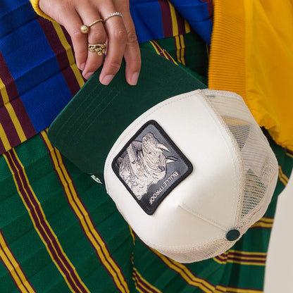 A person holds the Goorin Bros. Bulletproof Trucker cap in beige and green mesh with a rhino patch, wearing green striped pants and colorful clothing.