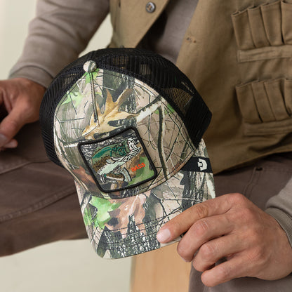 Person holding a Goorin Bros. Camouflage Bass trucker hat with mesh back, blending fishing fashion and outdoor style, featuring a bass patch and bold design.